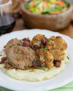 Pork Medallions & Cauliflower with Pinot Cherry Sauce over mashed potatoes on a white plate on a wooden serving tray with salad and red wine in the background