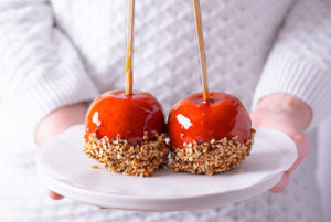 Person in a white sweater holding out a white tray with Caramel Apples dipped in crushed hazelnuts