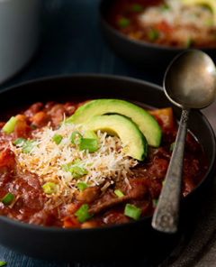 Vegetarian Chili topped with avocado slices and green onions in a black bowl on a dark blue moody background