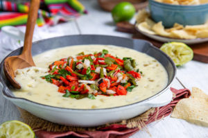Queso in a cast iron skillet topped with red and green peppers for Christmas and a wooden spoon resting in the skillet