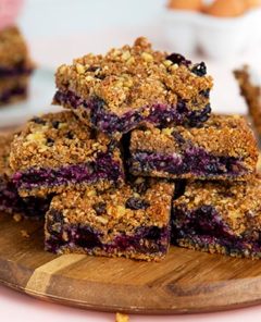 Granola-Topped Blueberry Pie Bars stacked on a round wooden cutting board on a light pink background