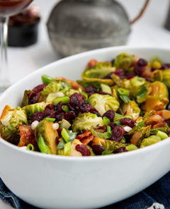 Soy-Glazed Brussels Sprouts in a white oval serving dish with glasses of red wine in the background
